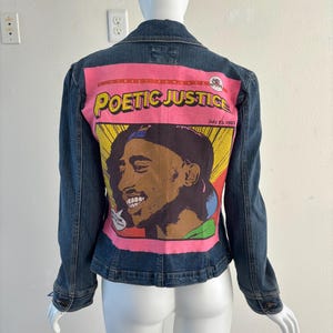 May include: A denim jacket with a graphic on the back featuring the words "POETIC JUSTICE" and an illustration of a person. The graphic is set against a pink background with yellow and red accents. The jacket is displayed on a mannequin.