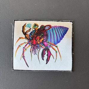 May include: A colorful embroidered patch featuring a detailed illustration of a crustacean-like creature with a shell. The creature has multiple eyes, claws, and legs, and is adorned with a gem. The shell is striped with blue, purple, and pink.
