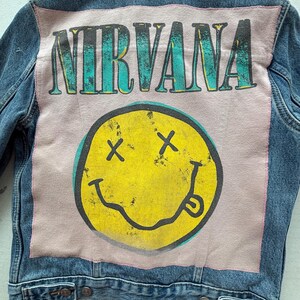 May include: A denim jacket with a large graphic on the back. The graphic features the word "NIRVANA" in teal letters above a yellow smiley face with black cross eyes and a tongue sticking out. The jacket is blue.