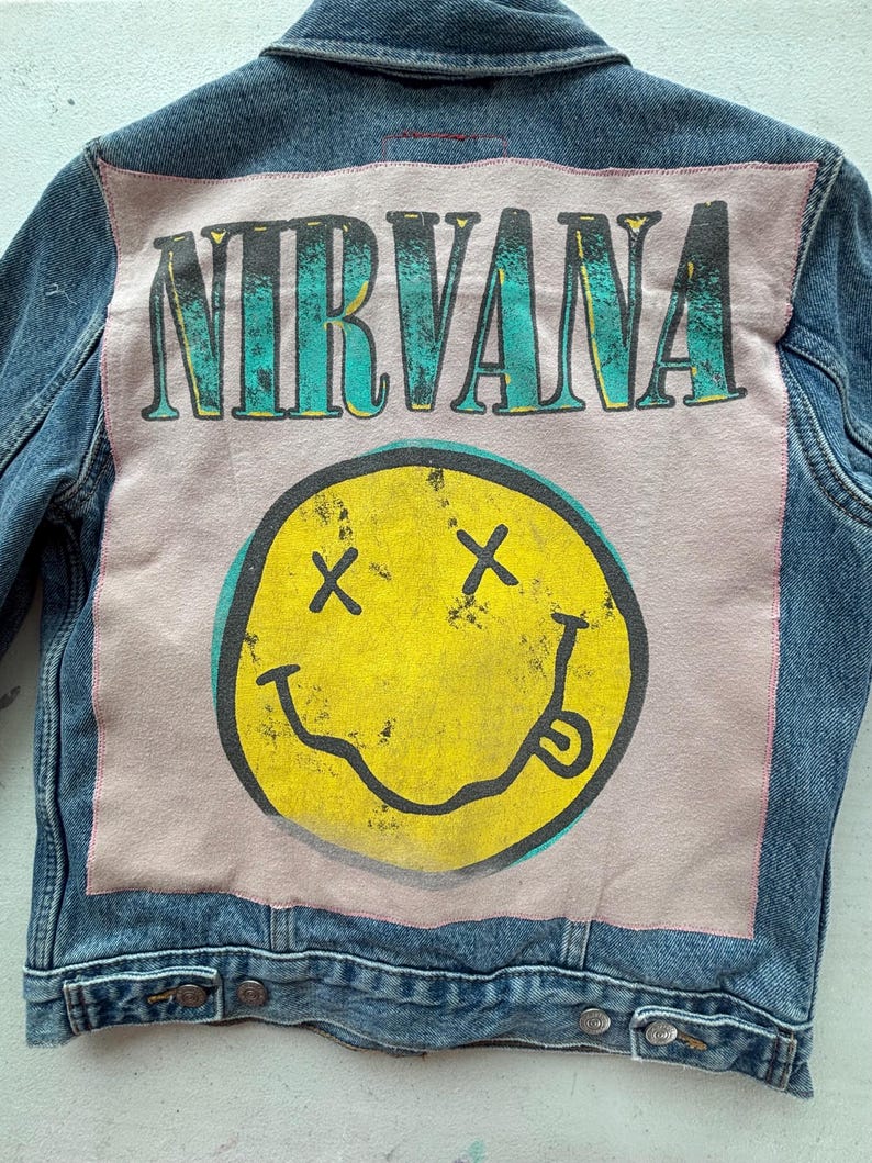 May include: A denim jacket with a large graphic on the back. The graphic features the word "NIRVANA" in teal letters above a yellow smiley face with black cross eyes and a tongue sticking out. The jacket is blue.