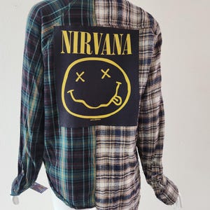 May include: A long-sleeved plaid shirt in shades of green, blue, and cream. The back features a large black patch with the word "NIRVANA" in yellow above a yellow smiley face with crossed-out eyes and a protruding tongue. The shirt is displayed on a mannequin.