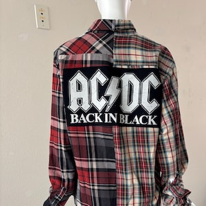 May include: A long-sleeved plaid shirt in red, black, and cream. The back features a large black patch with the AC/DC logo and the text "BACK IN BLACK" in white. The shirt is displayed on a mannequin.