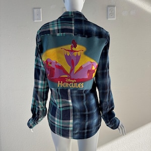 May include: A blue and green plaid flannel shirt with a graphic of Disney's Hercules on the back. The graphic features a character in a purple dress with yellow and red accents, and the text "Disney's Hercules".