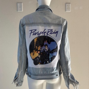 May include: Light blue denim jacket with a graphic on the back featuring the words "Purple Rain" and an image of a person on a motorcycle. The jacket has long sleeves, a button-down front, and two chest pockets.