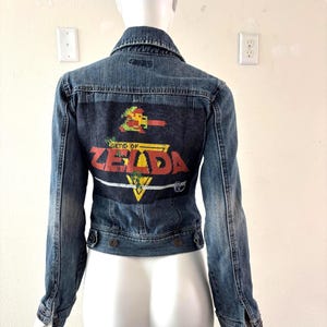 May include: A blue denim jacket with a graphic on the back featuring the words "Legend of Zelda" in red and yellow. The jacket has a classic collar, button closures, and long sleeves.