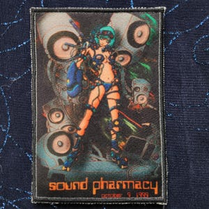 May include: A rectangular patch featuring a futuristic anime-style illustration. The image depicts a female figure in a blue outfit with a weapon, surrounded by speakers and machinery. The text "sound pharmacy october 9 1999" is printed at the bottom.