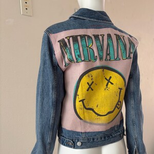 May include: A blue denim jacket with a light pink back panel featuring the word "NIRVANA" in teal lettering and a yellow smiley face with crossed-out eyes. The jacket has a classic button-up design.
