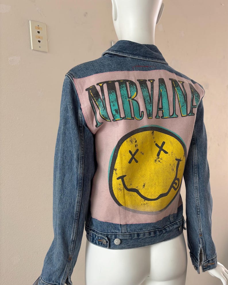 May include: A blue denim jacket with a light pink back panel featuring the word "NIRVANA" in teal lettering and a yellow smiley face with crossed-out eyes. The jacket has a classic button-up design.