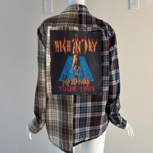 May include: A plaid flannel shirt with a large patch on the back featuring the text "HIGH N' DRY" and "DEF LEPPARD TOUR 1981". The shirt is a mix of brown, gray, and blue plaid patterns.