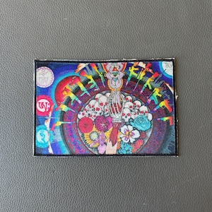 May include: A rectangular patch with a vibrant, psychedelic design. The artwork features a central arrangement of skulls, flowers, and a skeletal hand, surrounded by colorful circles, guns, and Tibetan script. The color palette includes blues, reds, yellows, and greens.