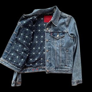 May include: A denim jacket with a light blue wash. The jacket's interior lining features a dark blue fabric with a white geometric starburst pattern. The jacket has a red tag and red buttons on the cuffs and bottom hem.