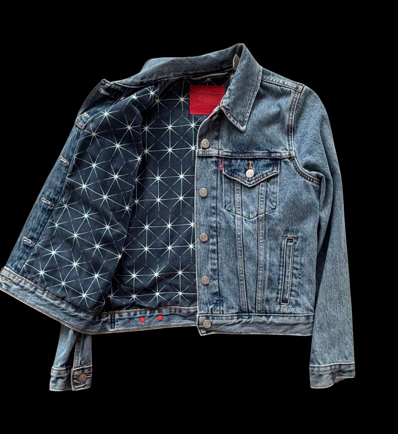 May include: A denim jacket with a light blue wash. The jacket's interior lining features a dark blue fabric with a white geometric starburst pattern. The jacket has a red tag and red buttons on the cuffs and bottom hem.