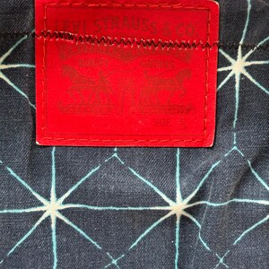 May include: Close-up of a red Levi Strauss & Co. label stitched onto a dark blue denim fabric with a white and light blue geometric pattern. The label features the brand name and logo.