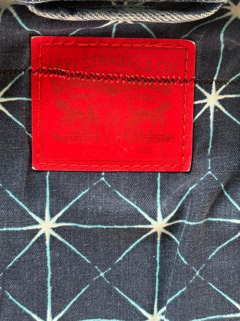 May include: Close-up of a red Levi Strauss & Co. label stitched onto a dark blue denim fabric with a white and light blue geometric pattern. The label features the brand name and logo.