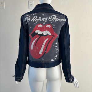 May include: Dark blue denim jacket with a graphic of the Rolling Stones' iconic tongue and lips logo on the back. The logo is red and white against a gray background, with the band's name in white script above. The jacket has a classic design.