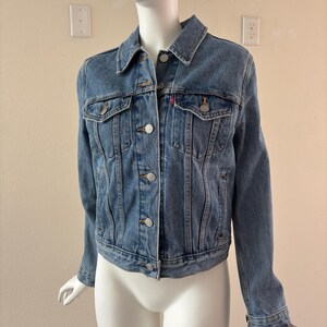 May include: A light blue denim jacket with a classic design. Features a button-up front, two chest pockets, and a small red logo tag. The jacket is displayed on a mannequin.