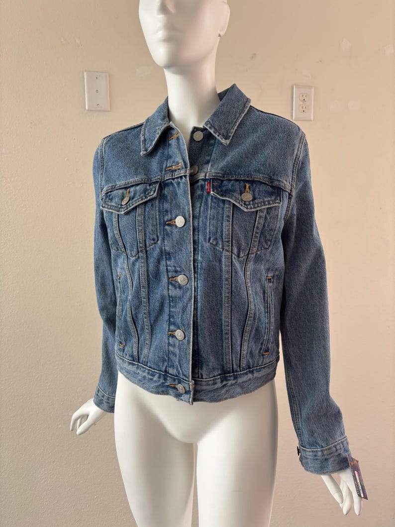 May include: A light blue denim jacket with a classic design. Features a button-up front, two chest pockets, and a small red logo tag. The jacket is displayed on a mannequin.