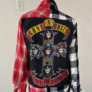 May include: A long-sleeved shirt with a Guns N' Roses graphic on the back. The shirt features a patchwork design with red plaid on the left side and black and white plaid on the right. The graphic includes skulls and the words "Appetite for Destruction."