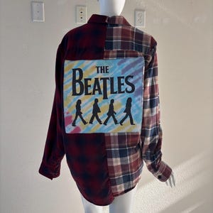 May include: A flannel shirt with a patchwork design. The left side is a dark red plaid, and the right side is a burgundy, navy, and white plaid. The back features a tie-dye design with "The Beatles" and the band's silhouette.