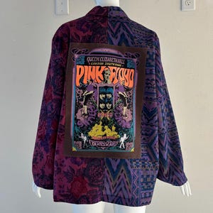 May include: A patchwork jacket with a large Pink Floyd concert poster on the back. The jacket features a mix of purple floral and geometric patterns. The poster is brown with orange, purple, and blue text and images.