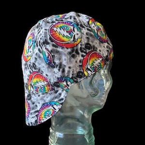 May include: A gray and white patterned welding cap featuring colorful tie-dye skull and lightning bolt designs. The cap is made of fabric and has a curved brim. The background is black.