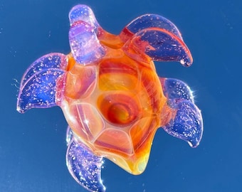 May include: A colorful glass turtle figurine with a vibrant blend of orange, yellow, and red hues on its shell. The flippers and head are a translucent blue, creating a striking contrast. The turtle is a decorative item.