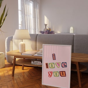 May include: A framed print with a pink background and white frame. The print features the words "i love you" in colorful, cut-out letters.