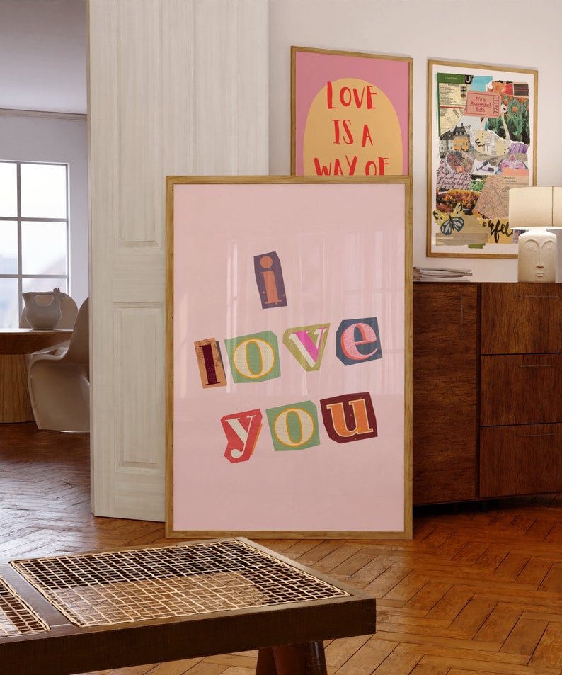 May include: A pink framed poster with the words "i love you" in colorful, cut-out letters. The letters are arranged in a staggered pattern. The poster is hanging on a wall in a room with wooden floors.