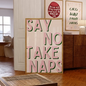 Op de afbeelding: Een ingelijste poster met de tekst "SAY NO TAKE NAPS" in roze letters met groene contouren. Twee andere ingelijste posters zijn zichtbaar, een met de tekst "GIRLS WANT DINNER PARTIES" en een andere met Franse tekst. De posters worden in een kamer met houten vloeren getoond.