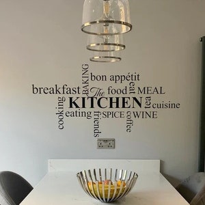 May include: A white kitchen table with a black and white word art decal that says "The Kitchen" with words like "baking", "breakfast", "bon appétit", "cooking", "eating", "friends", "food", "meal", "spice", "wine", and "coffee" surrounding the main text. The table has a fruit bowl in the centre.