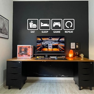 May include: A gaming desk setup featuring a black wall with a white vinyl decal displaying the text EAT SLEEP GAME REPEAT with corresponding icons. The desk has two black drawer units, a computer tower, a monitor, a keyboard, and a mouse.