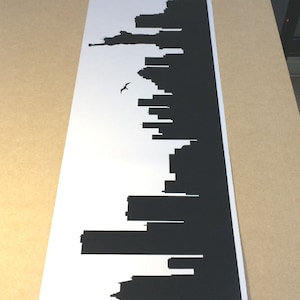 May include: A long, rectangular white decal featuring a black silhouette of the New York City skyline. The design includes various skyscrapers, the Statue of Liberty, and a single bird in flight, all rendered in a solid, high-contrast black graphic.