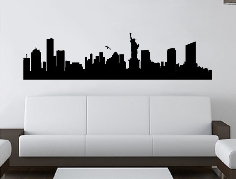 May include: A black wall decal featuring a detailed silhouette of the New York City skyline, including various skyscrapers and the Statue of Liberty. A small bird is depicted in flight above the buildings, creating a wide, horizontal graphic.