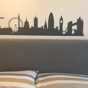 May include: A dark gray wall decal featuring a silhouette of the London skyline, including the London Eye, Big Ben, and Tower Bridge, with a small airplane flying above the city structures, applied to a plain, light-colored wall surface.