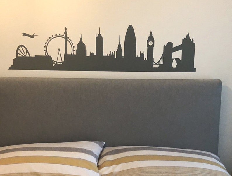 May include: A dark gray wall decal featuring a silhouette of the London skyline, including the London Eye, Big Ben, and Tower Bridge, with a small airplane flying above the city structures, applied to a plain, light-colored wall surface.