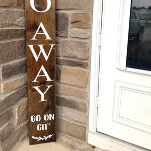 May include: A brown wooden sign with white lettering that reads "GO AWAY GO ON GIT". The sign is decorated with small white floral designs at the top and bottom.