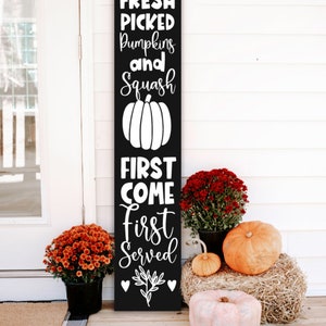 May include: Black wooden sign with white text that reads "Fresh Picked Pumpkins and Squash First Come First Served" with a white pumpkin graphic. The sign is leaning against a white wooden wall with fall decorations.