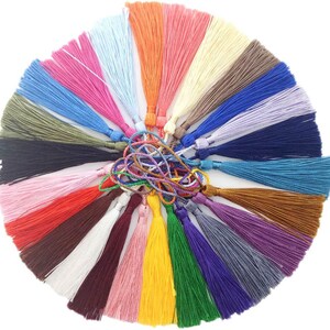 K&ouml;nnte beinhalten: Eine Sammlung von 24 bunten Quasten, jede in einer anderen Farbe, darunter Rot, Blau, Gr&uuml;n, Gelb, Lila, Orange, Braun, Schwarz, Wei&szlig; und Rosa. Die Quasten sind in einem Kreis angeordnet, wobei die Spitzen der Quasten nach au&szlig;en zeigen.