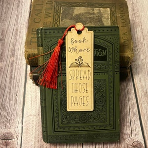 K&ouml;nnte beinhalten: Ein Holz-Lesezeichen mit einem roten Quasten. Das Lesezeichen hat den Text "Book Whore Spread Those Pages" und eine Illustration eines offenen Buches mit Blumen.