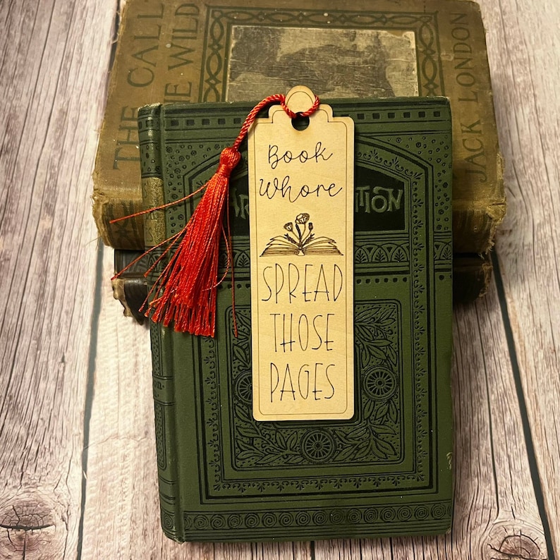 K&ouml;nnte beinhalten: Ein Holz-Lesezeichen mit einem roten Quasten. Das Lesezeichen hat den Text "Book Whore Spread Those Pages" und eine Illustration eines offenen Buches mit Blumen.