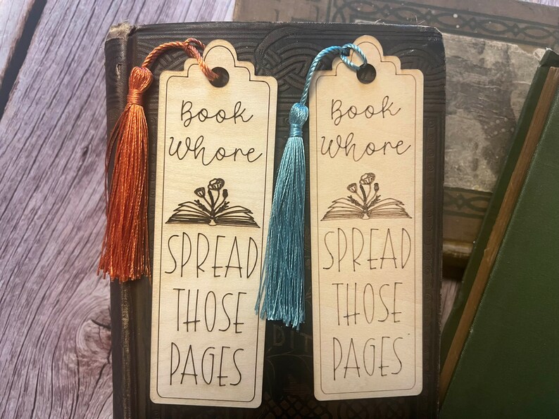 K&ouml;nnte beinhalten: Zwei Holzbuchzeichen mit einem Buch- und Blumendesign. Die Lesezeichen haben den Text "Book Whore Spread Those Pages" und werden mit einem Quasten geliefert. Ein Lesezeichen hat eine blaue Quaste und das andere eine orangefarbene Quaste.