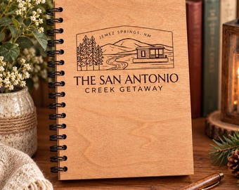 May include: A spiral-bound notebook with a light brown wooden cover featuring a black laser-engraved graphic of a cabin, trees, and mountains. The text reads JEMEZ SPRINGS, NM, THE SAN ANTONIO, and CREEK GETAWAY in a clean, sans-serif font.