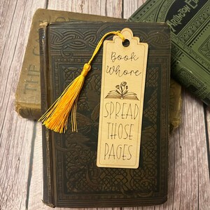 K&ouml;nnte beinhalten: Ein braunes Holz-Lesezeichen mit einem gelben Quasten. Das Lesezeichen hat den Text "Book Whore Spread Those Pages" und eine Illustration eines offenen Buches mit Blumen.