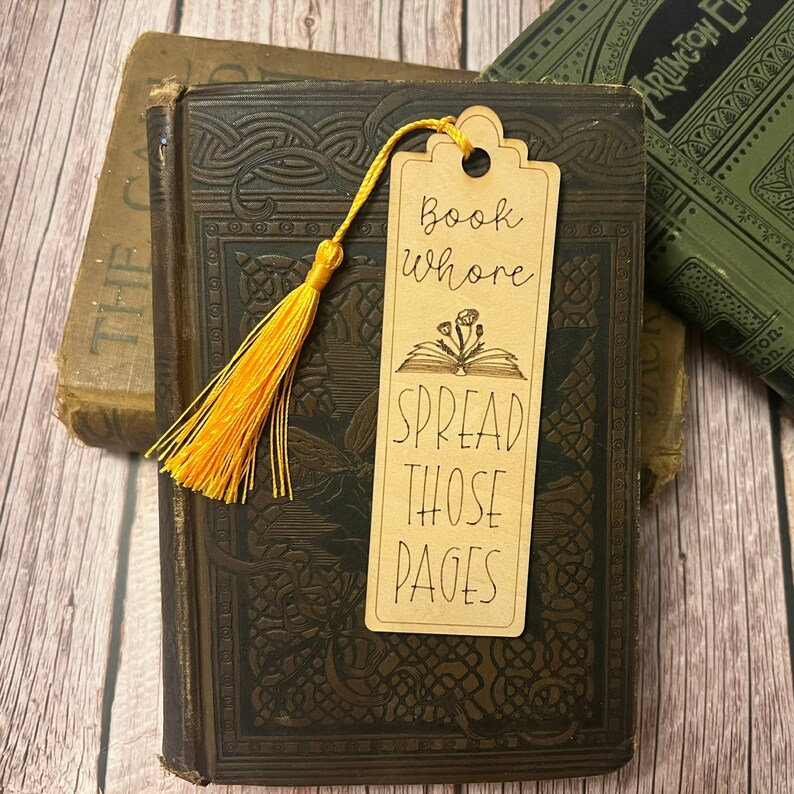 K&ouml;nnte beinhalten: Ein braunes Holz-Lesezeichen mit einem gelben Quasten. Das Lesezeichen hat den Text "Book Whore Spread Those Pages" und eine Illustration eines offenen Buches mit Blumen.
