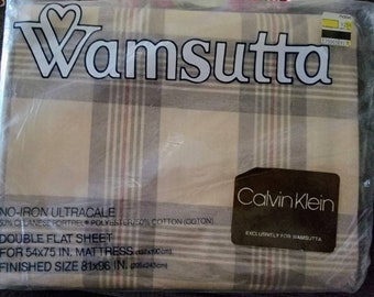 May include: A packaged Wamsutta double flat sheet with a plaid pattern in beige, gray, and red. The packaging includes the Calvin Klein logo and text indicating the sheet is for a 54x75 inch mattress, with a finished size of 81x96 inches.