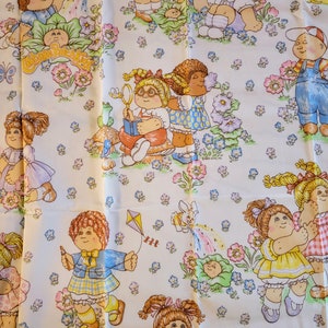 May include: A white fabric with a repeating pattern of Cabbage Patch Kids dolls. The dolls are wearing various outfits and are surrounded by flowers and butterflies. The text "Cabbage Patch Kids" is printed in the center of the fabric.