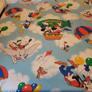 May include: A blue and white bed sheet with a Disney theme. The sheet features Mickey Mouse, Minnie Mouse, Donald Duck, Goofy, Dumbo, and Chip and Dale. The characters are flying in hot air balloons, parachutes, and airplanes. The sheet has a light blue background with white clouds.