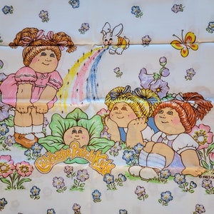May include: A white fabric with a Cabbage Patch Kids design. Three Cabbage Patch Kids dolls are smiling and looking up at a rainbow. The dolls are surrounded by flowers and a white bunny.