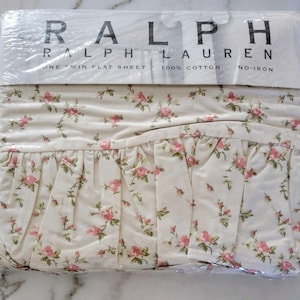 May include: A white twin flat sheet with a pink rose floral pattern. The sheet is packaged in plastic and has a label that reads "Ralph Lauren, One Twin Flat Sheet, 100% Cotton, No-Iron."