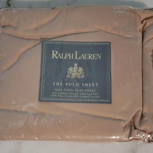May include: A new, unopened package of Ralph Lauren "The Polo Sheet" in a neutral tan color. The package includes a twin flat sheet made of 100% cotton with a 250 thread count. The label features the Ralph Lauren logo and product information.